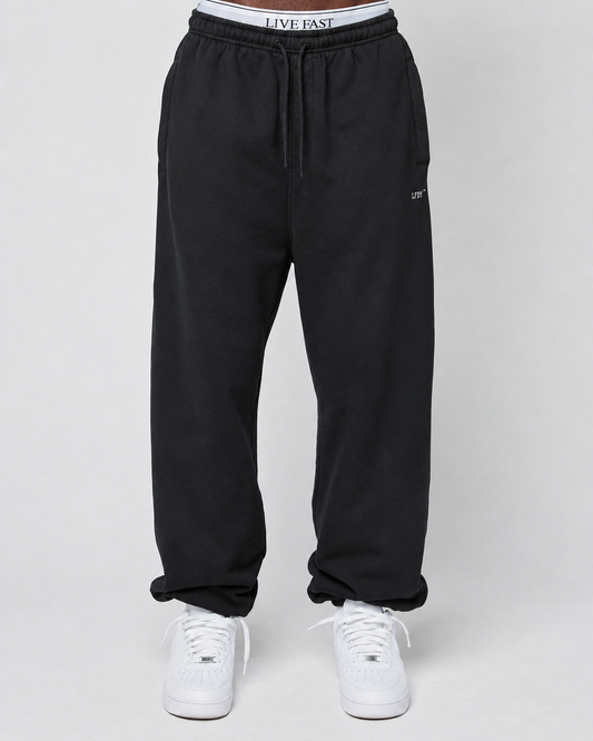 A person wears the Basic Loose Jogger by LIVE FAST DIE YOUNG—black, loose-fit sweatpants with an elastic waistband featuring “LIVE FAST” text, paired with white sneakers, against a plain light background.