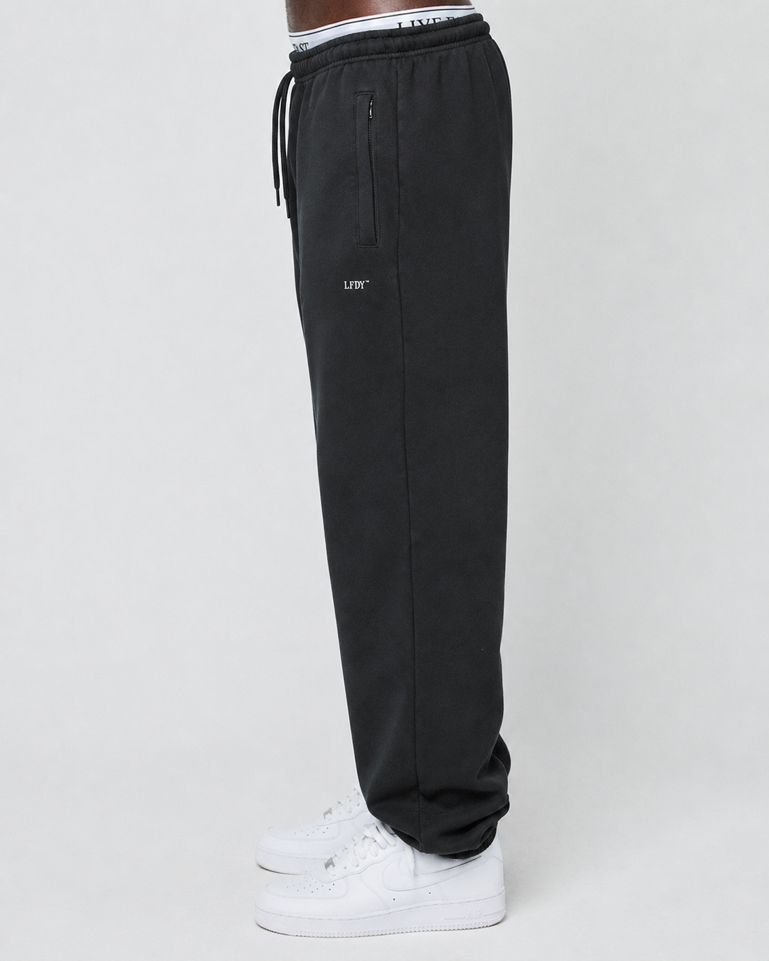 A person stands sideways against a plain light background wearing LIVE FAST DIE YOUNG Basic Loose Jogger pants with a loose fit, Elastik-Bund waistband, zipper pocket, white inner lining with text, and white sneakers.