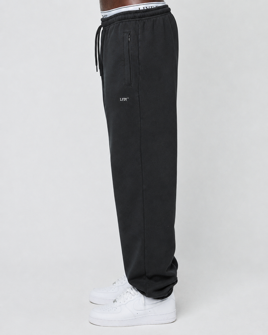 A person stands sideways against a plain light background wearing LIVE FAST DIE YOUNG Basic Loose Jogger pants with a loose fit, Elastik-Bund waistband, zipper pocket, white inner lining with text, and white sneakers.