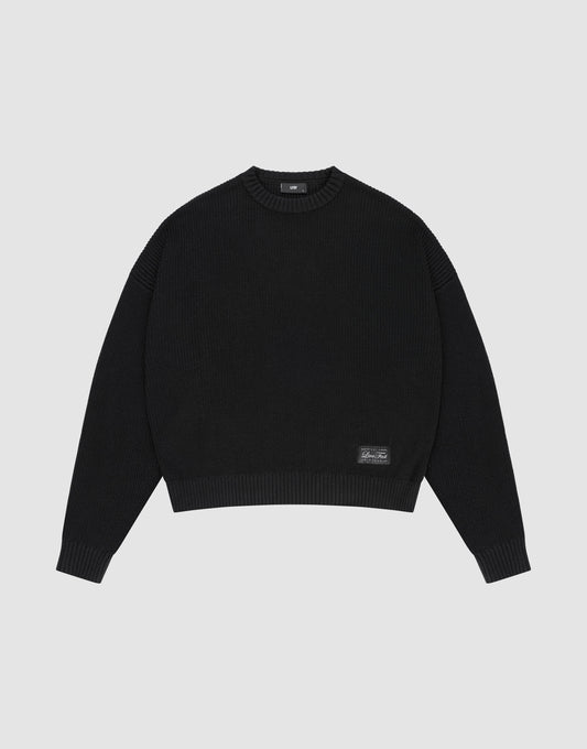 The LFDY Knit Sweater by LIVE FAST DIE YOUNG is a black, long-sleeve, oversized knit with dropped shoulders, crew neck, ribbed cuffs and hem, and a small rectangular patch near the bottom left hem, shown on a plain light gray background. | plain