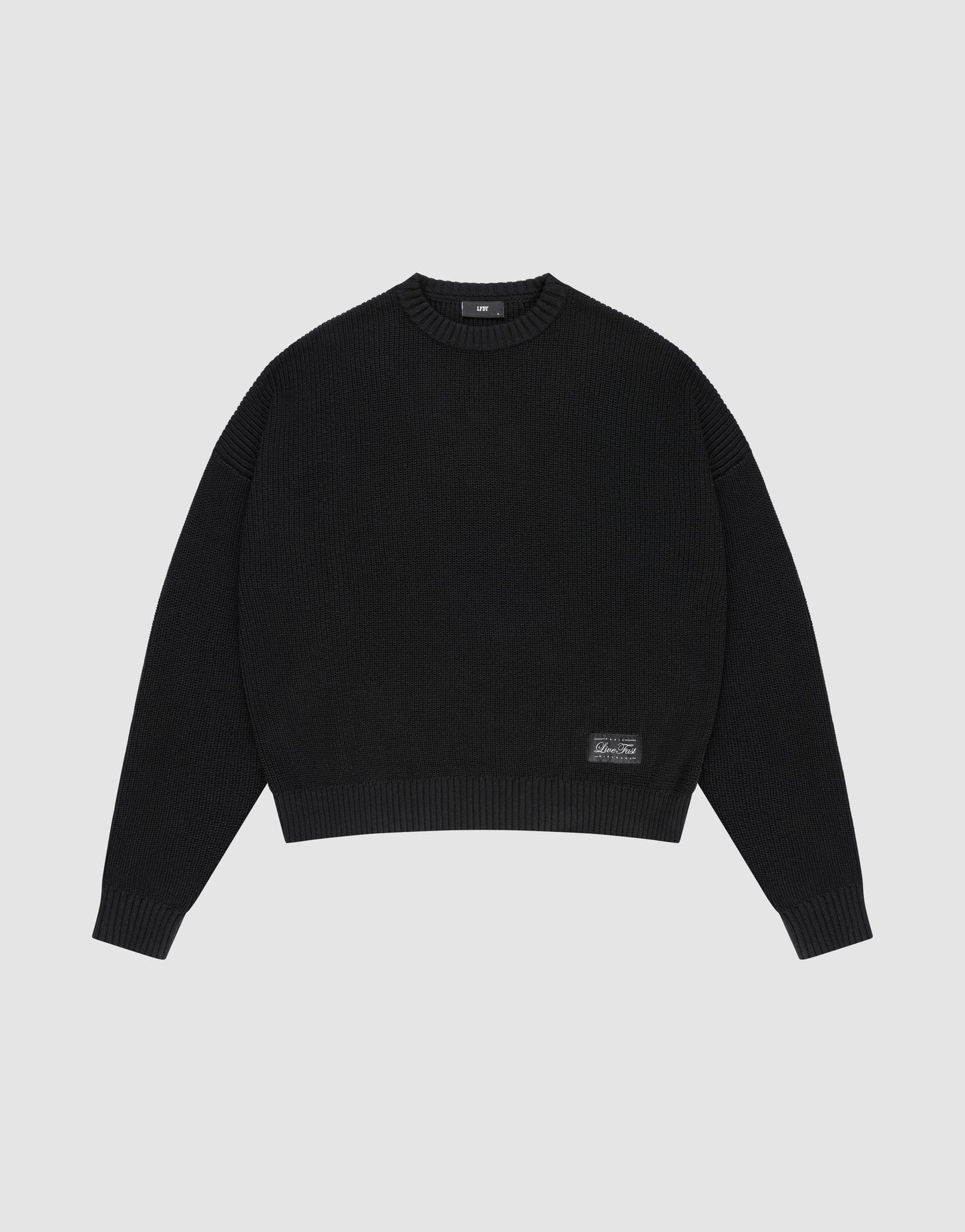 The LFDY Knit Sweater by LIVE FAST DIE YOUNG is a black, long-sleeve, oversized knit with dropped shoulders, crew neck, ribbed cuffs and hem, and a small rectangular patch near the bottom left hem, shown on a plain light gray background. | plain