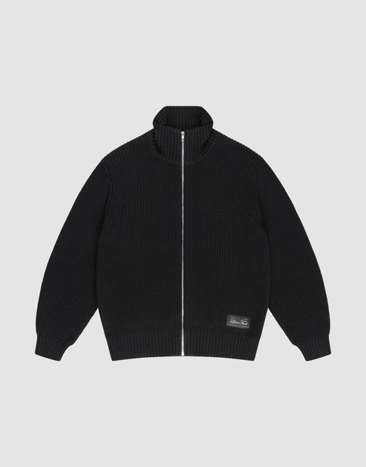The LFDY Knit Jacket by LIVE FAST DIE YOUNG is a black, ribbed zip-up with Stehkragen, long sleeves, and a small rectangular patch near the hem. Made from 100% Baumwolle, it's shown laid flat on a light gray background. | plain