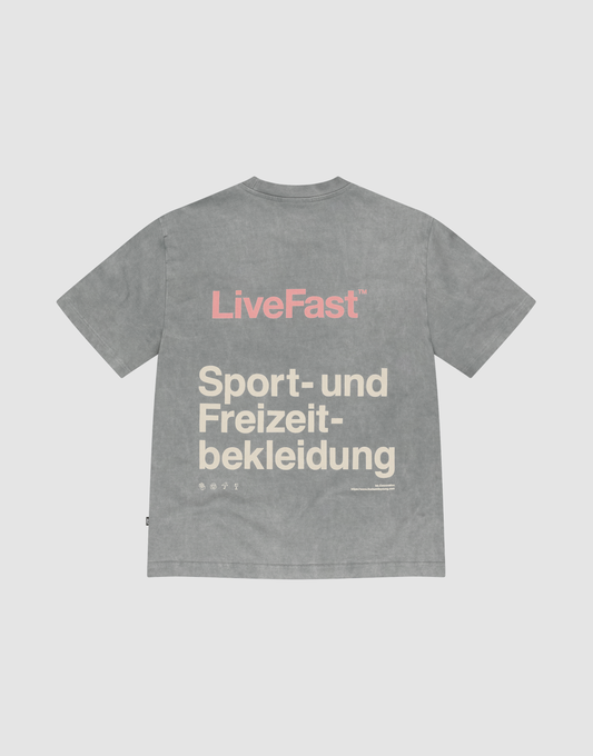 The LIVE FAST DIE YOUNG Basic Trademark Tee is a gray, short-sleeve, oversize-fit t-shirt made from 100% Baumwolle. It features "LiveFast" in pink and “Sport- und Freizeitbekleidung” in beige on the back, shown laid flat on a light background.