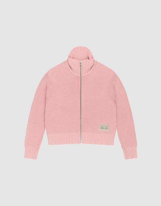 The LFDY Knit Jacket by LIVE FAST DIE YOUNG is a light pink, ribbed zip-up sweater with a stand collar and long sleeves, made from 100% cotton. A small beige patch accents the lower left front. Shown here laid flat on a white background. | plain