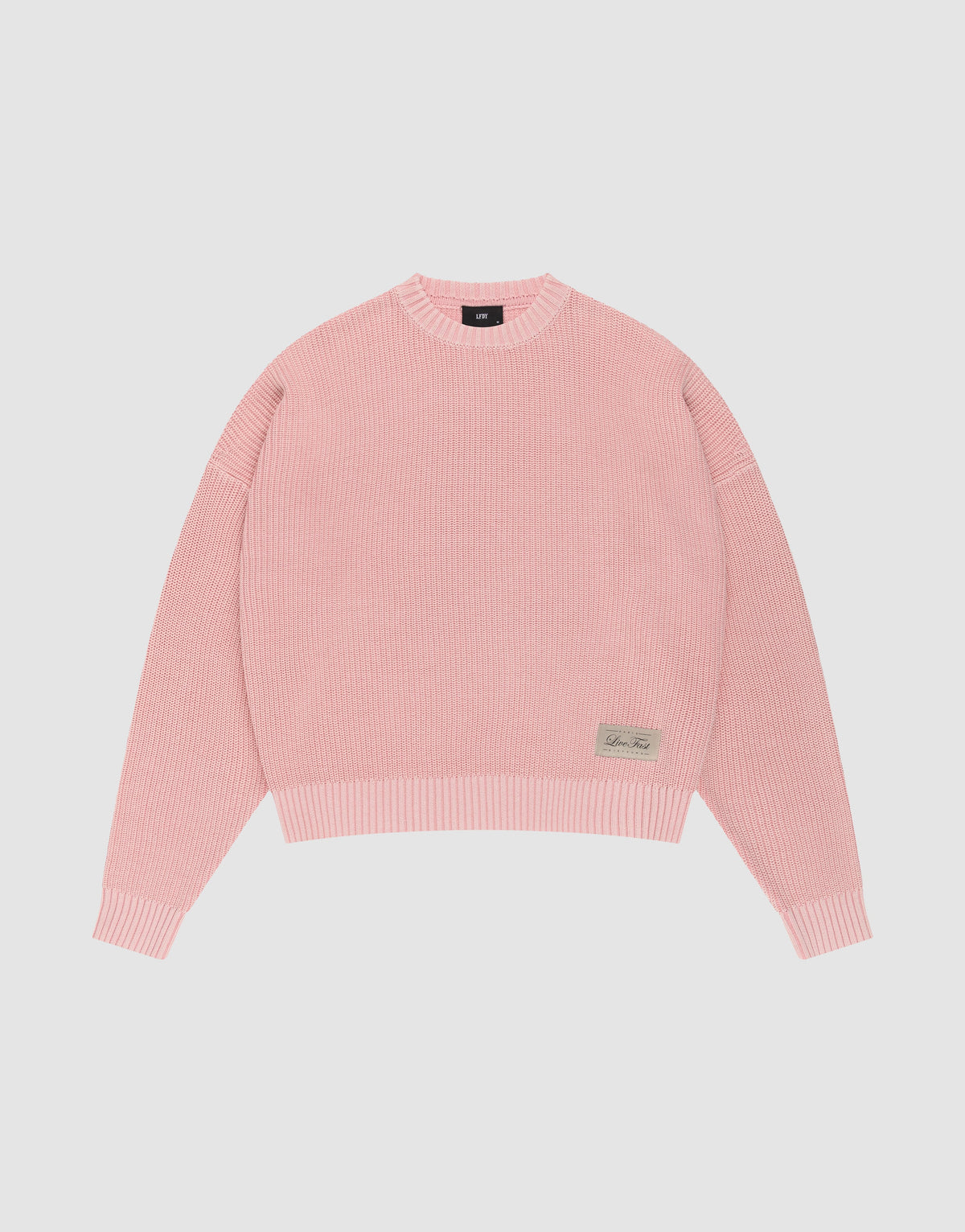The LFDY Knit Sweater by LIVE FAST DIE YOUNG features pink ribbed knit, an oversized fit, dropped shoulders, long sleeves, a crew neckline, and a small rectangular beige patch with text on the front hem, displayed on a plain white background. | plain