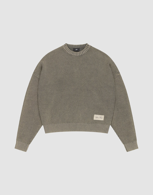 The LFDY Knit Sweater by LIVE FAST DIE YOUNG is a ribbed, oversized crewneck in muted olive green Baumwolle with long sleeves, dropped shoulders, and a small cream text patch on the front left. Shown flat on a light gray background. | plain