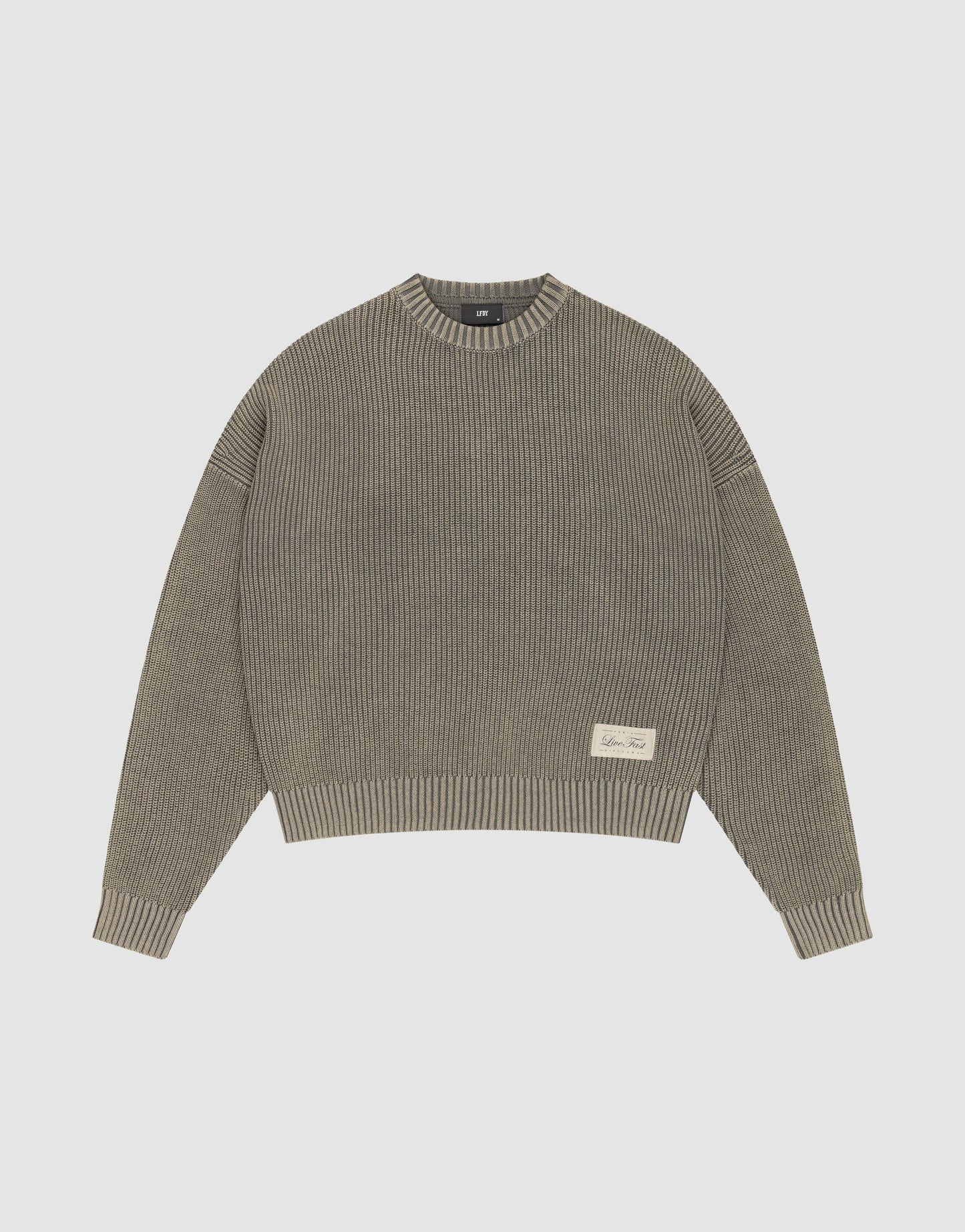 The LFDY Knit Sweater by LIVE FAST DIE YOUNG is a ribbed, oversized crewneck in muted olive green Baumwolle with long sleeves, dropped shoulders, and a small cream text patch on the front left. Shown flat on a light gray background. | plain