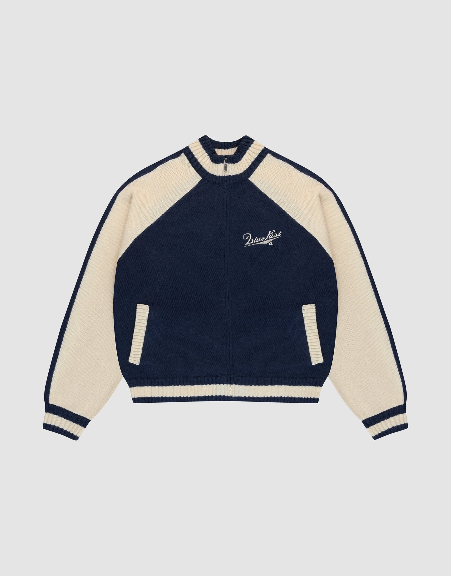 The LIVE FAST DIE YOUNG Boxy Knit Zip Jacket features a navy and cream varsity style with cream raglan sleeves, striped cuffs and collar, two front pockets, and Paradise embroidery on the left chest. Set against a plain light gray background.