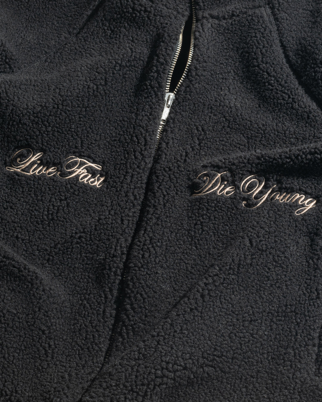 A close-up of the black LFDY Teddy Jacket by LIVE FAST DIE YOUNG, with raglan sleeves, a silver zipper, and "Live Fast" embroidered on the left and "Die Young" on the right in cursive.