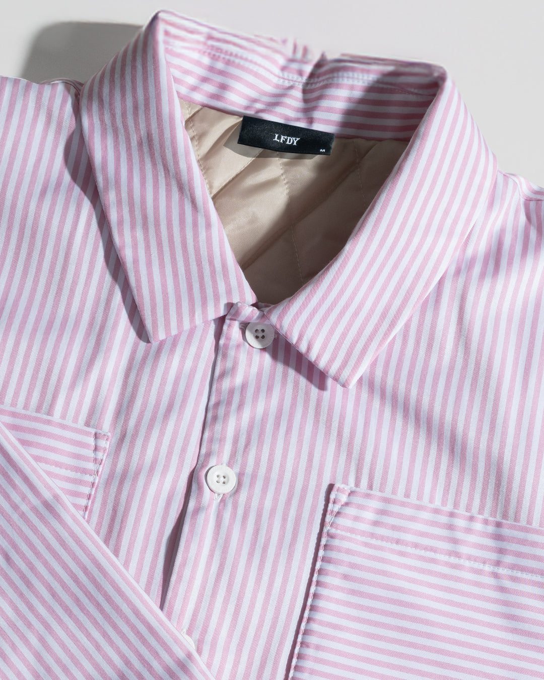 A close-up of the LIVE FAST DIE YOUNG LFDY Striped Padded Shirt in light pink and white stripes, featuring a folded collar, two pockets, and white buttons. This Herren Hemdjacke is neatly arranged on a light surface.
