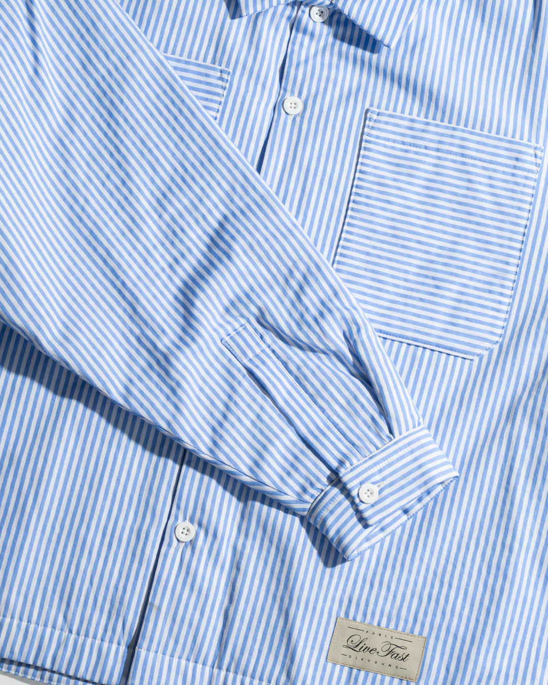The LIVE FAST DIE YOUNG LFDY Striped Padded Shirt features blue and white vertical stripes, a front pocket, white buttons, and a sewn label near the bottom hem.