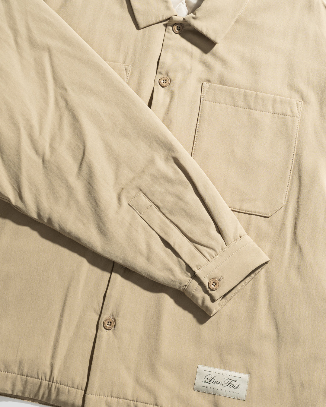 The LFDY Padded Shirt by LIVE FAST DIE YOUNG is a beige oversize overshirt with long sleeves, front chest pockets, light brown buttons, and a white rectangular Live Fast label sewn near the bottom hem.