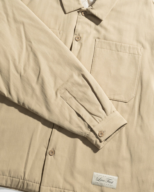 The LFDY Padded Shirt by LIVE FAST DIE YOUNG is a beige oversize overshirt with long sleeves, front chest pockets, light brown buttons, and a white rectangular Live Fast label sewn near the bottom hem.