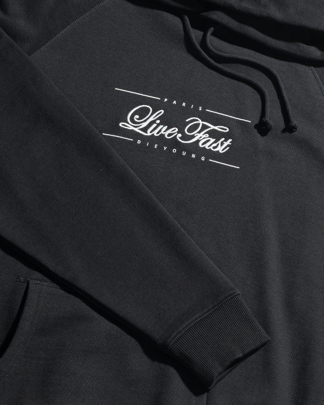 The LFDY Embroidery Hooded by LIVE FAST DIE YOUNG is a black Regular Fit Hoodie in soft 100% Baumwolle french terry, featuring drawstrings and white "PARIS Live Fast DIE YOUNG" text embroidered on the chest. Laid flat showing sleeve and pocket.