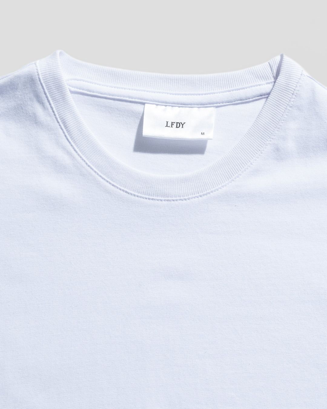 Close-up of the LIVE FAST DIE YOUNG LFDY Slim Tee in light blue, featuring a collar and white tag with LFDY and size M. Subtle flag label detail shown; made from 95% Baumwolle. Neutral light gray background.
