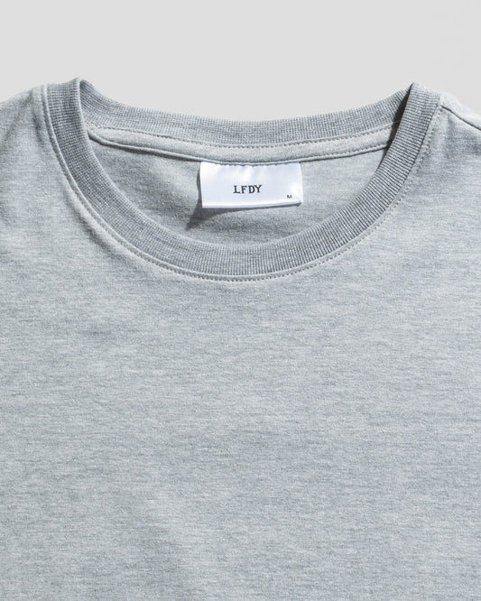 A close-up of the LFDY Slim Tee by LIVE FAST DIE YOUNG in light grey, featuring a white neck label with brand and size M, plus a signature Flag-Label. The slim fit T-shirt is shown laid flat on a light background.
