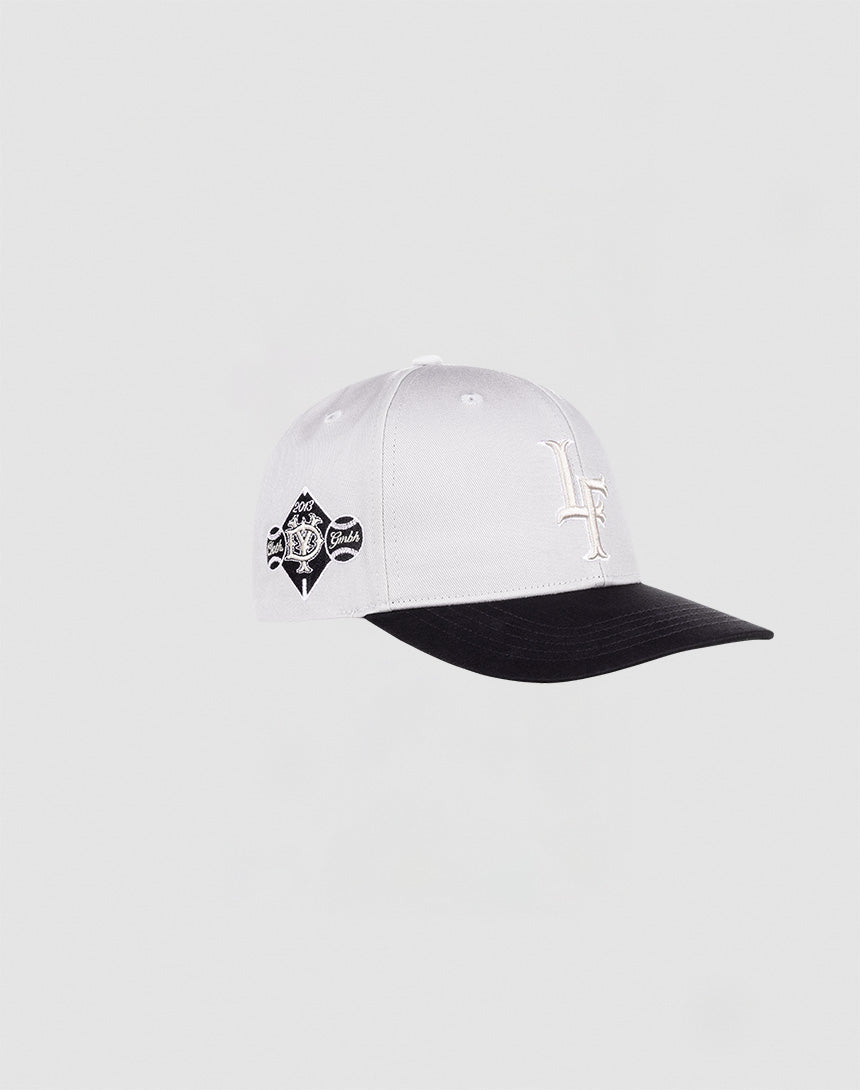 The LF Snapback by LIVE FAST DIE YOUNG is a white cap with a black curved brim and underside, front LF embroidered logo, black and white circular side patch, and snap closure, displayed against a plain light background.