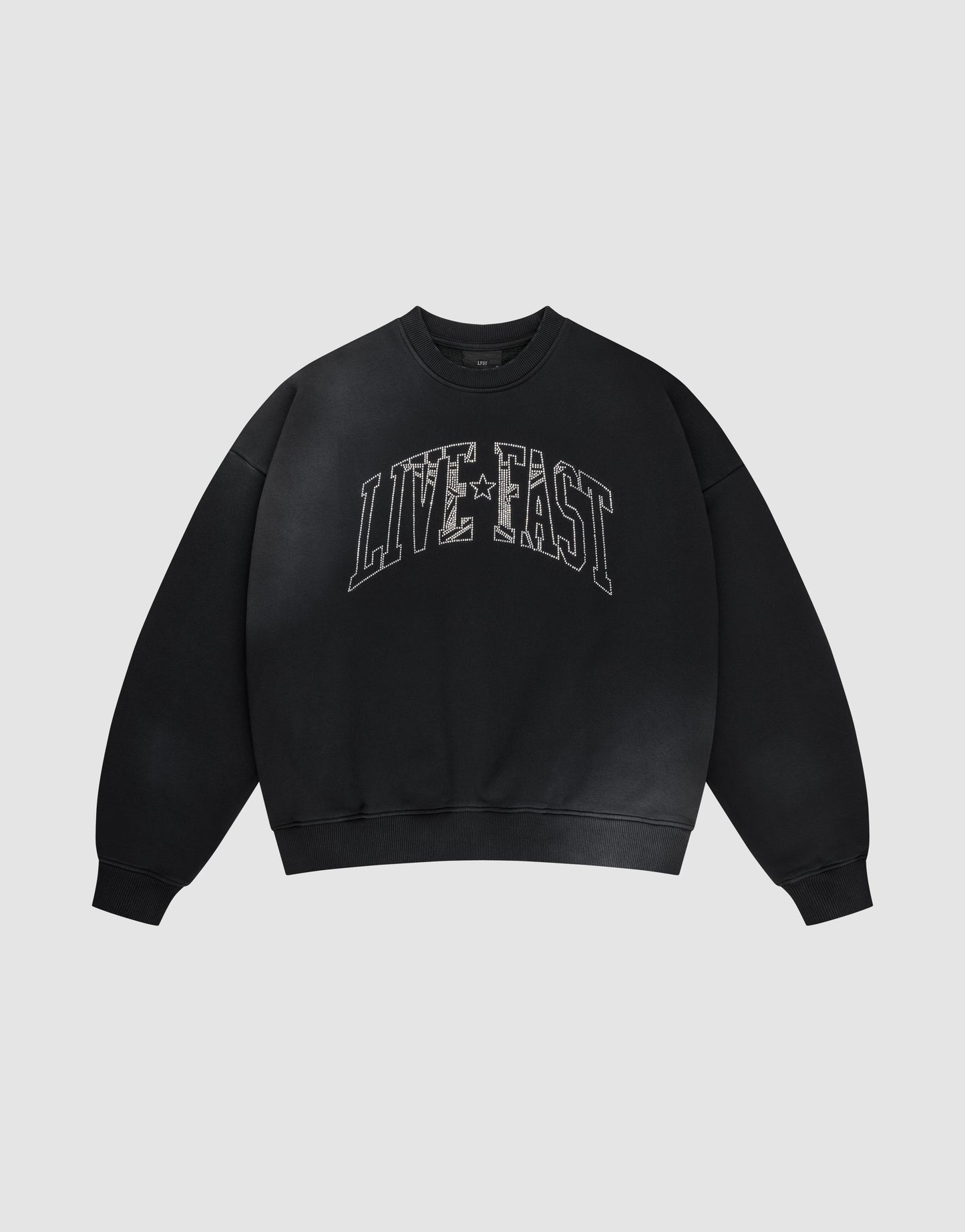 The LIVE FAST DIE YOUNG Live Fast Rhinestone Sweater is a black oversized crewneck with bold distressed white "LIVE FAST" lettering and a subtle spray effect, set against a plain light gray background.