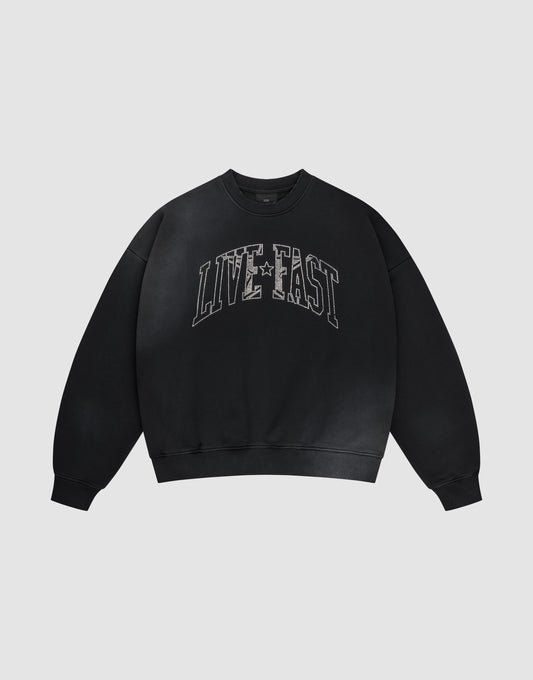 The LIVE FAST DIE YOUNG Live Fast Rhinestone Sweater is a black oversized crewneck with bold distressed white "LIVE FAST" lettering and a subtle spray effect, set against a plain light gray background. | plain