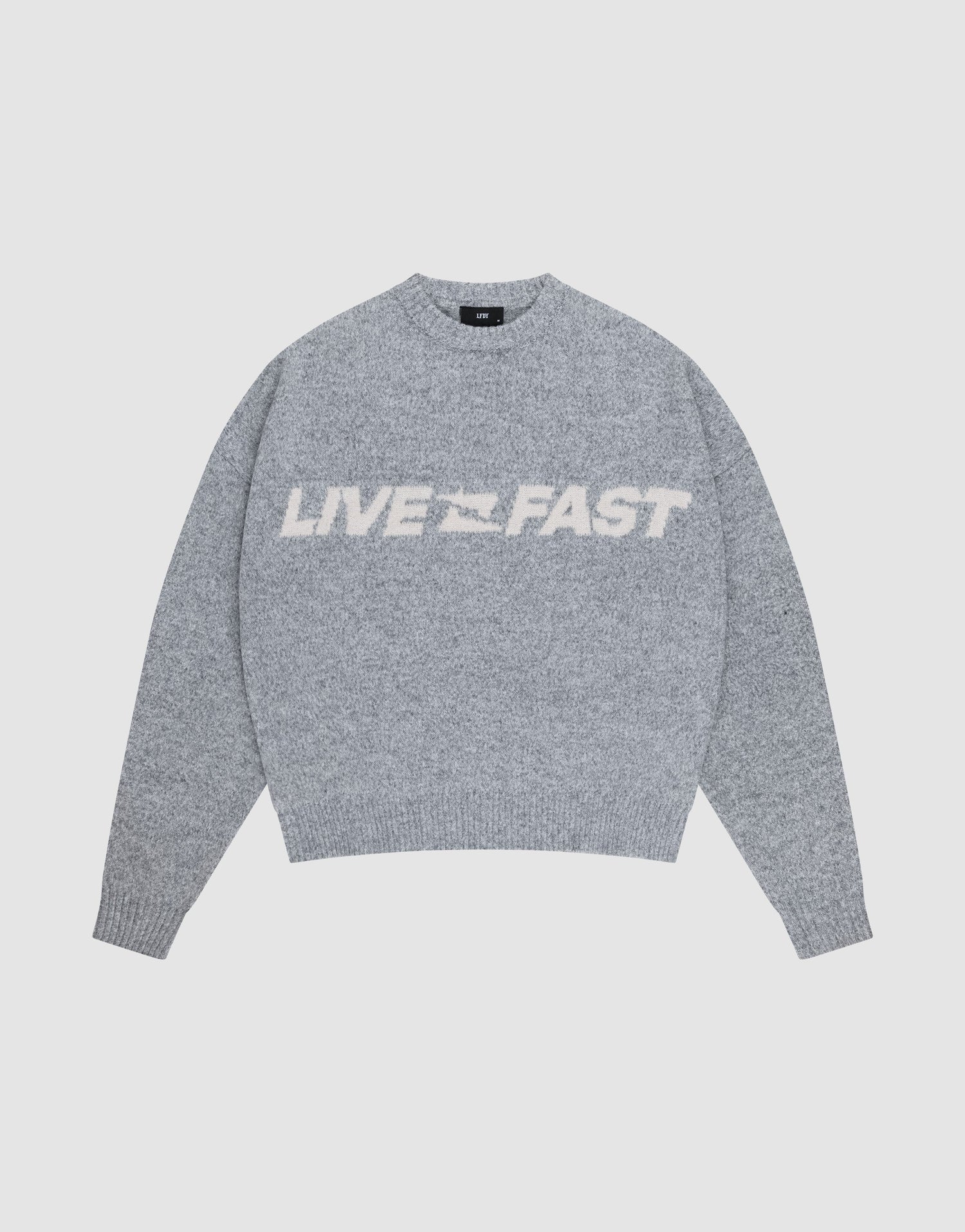 A person faces away, wearing an oversize light gray Live Fast Boucle Sweater and matching pants. The 100% polyester sweater by LIVE FAST DIE YOUNG displays LIVE FAST in bold white text across the back.