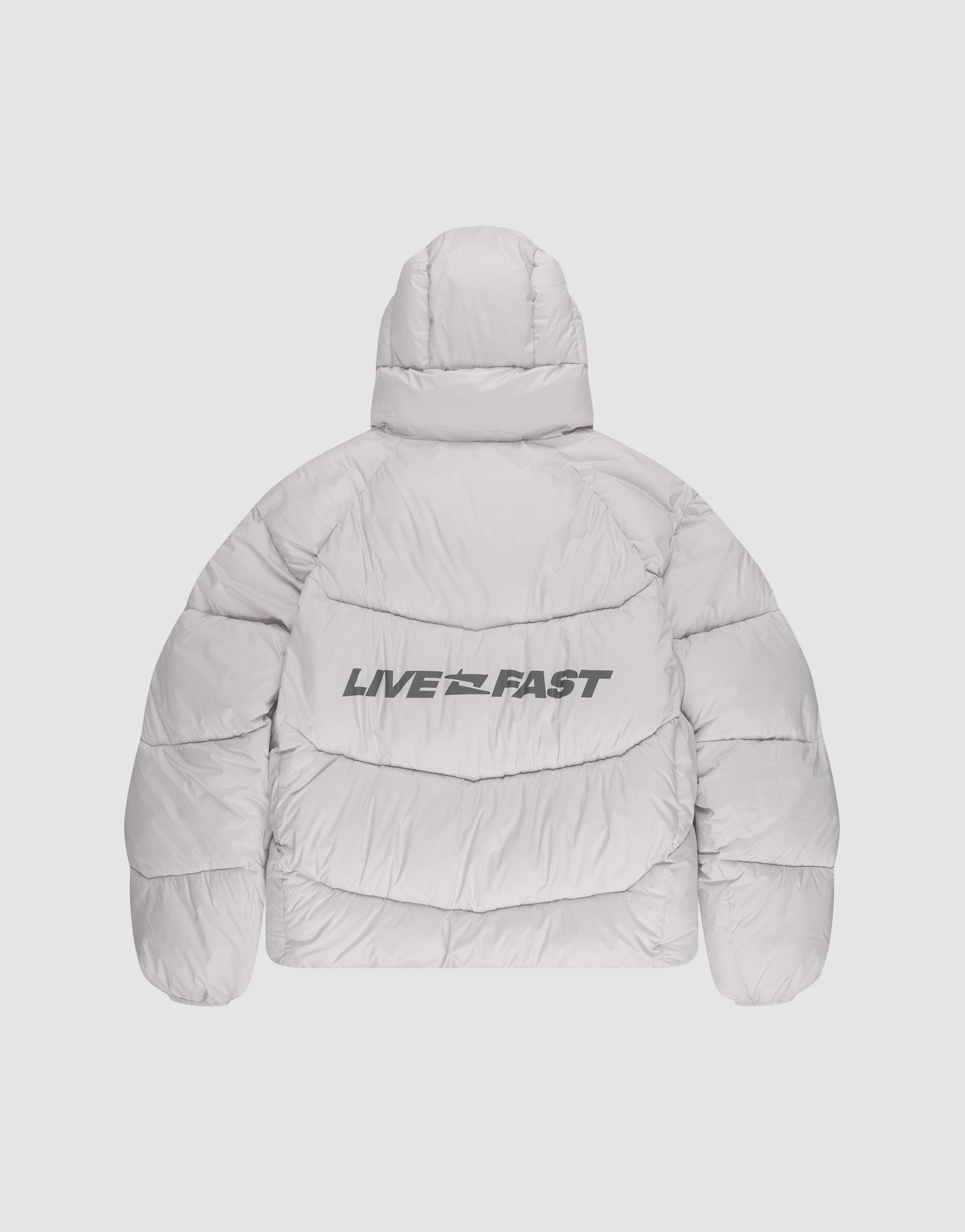 A person faces away, wearing the LIVE FAST DIE YOUNG Live Fast Puffer Jacket in oversize light gray with reflective "LIVE FAST" print on the back, black jeans, white shoes, and a matching beanie, set against a plain light background.
