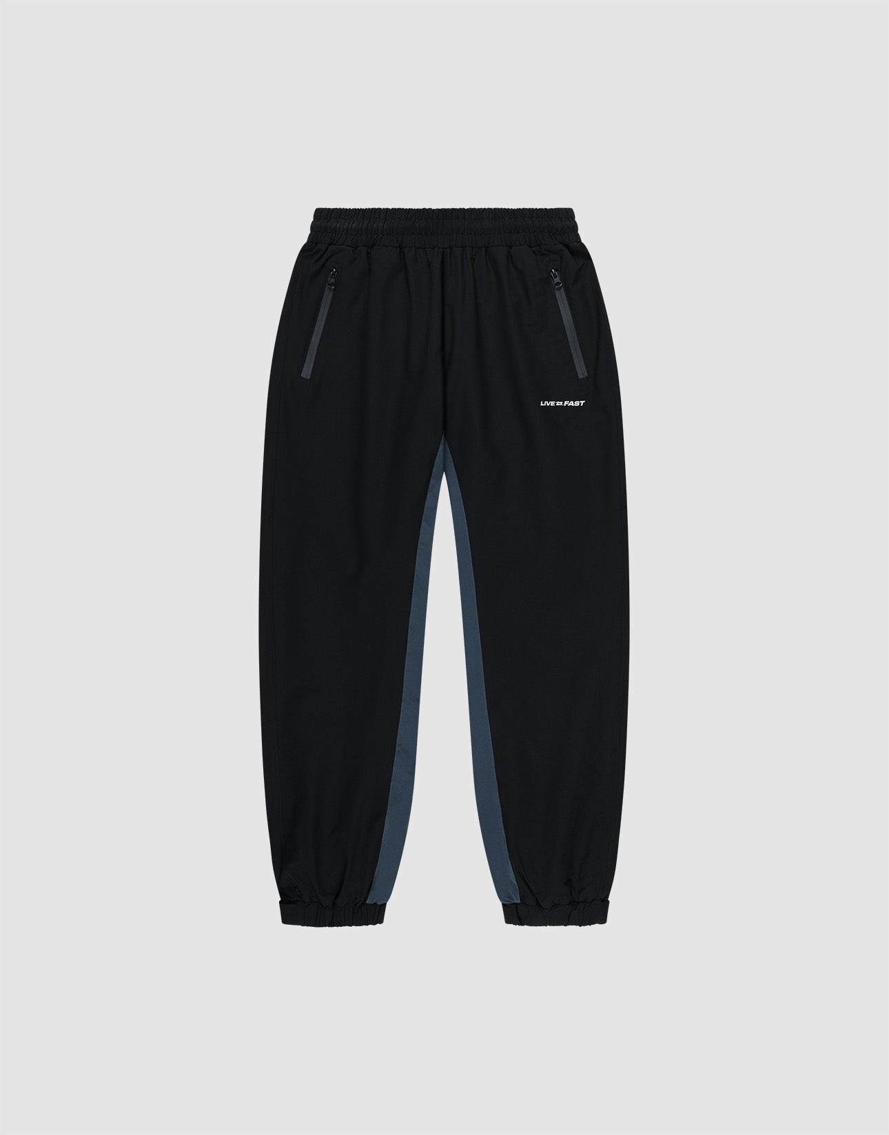 Black Basic Shell Pants by LIVE FAST DIE YOUNG with elastic waistband, zippered side pockets, gray panels, and elastic cuffs. Features a white reflective logo on the left thigh. Displayed on a plain light background.