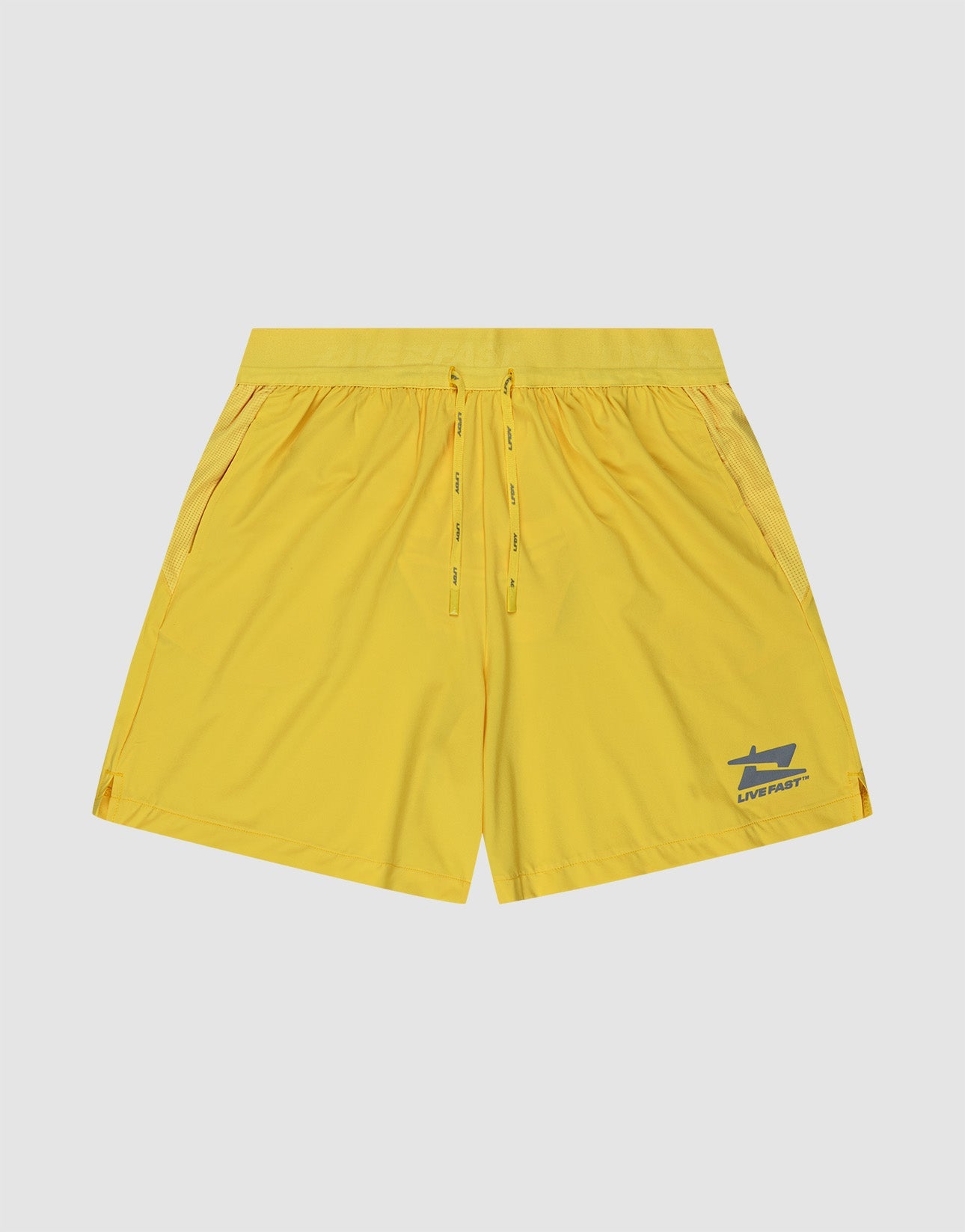 A person stands against a white background wearing LIVE FAST DIE YOUNG Live Fast Performance Shorts in bright yellow with a small left-leg logo and elastic drawstring waist, paired with white socks and sneakers.