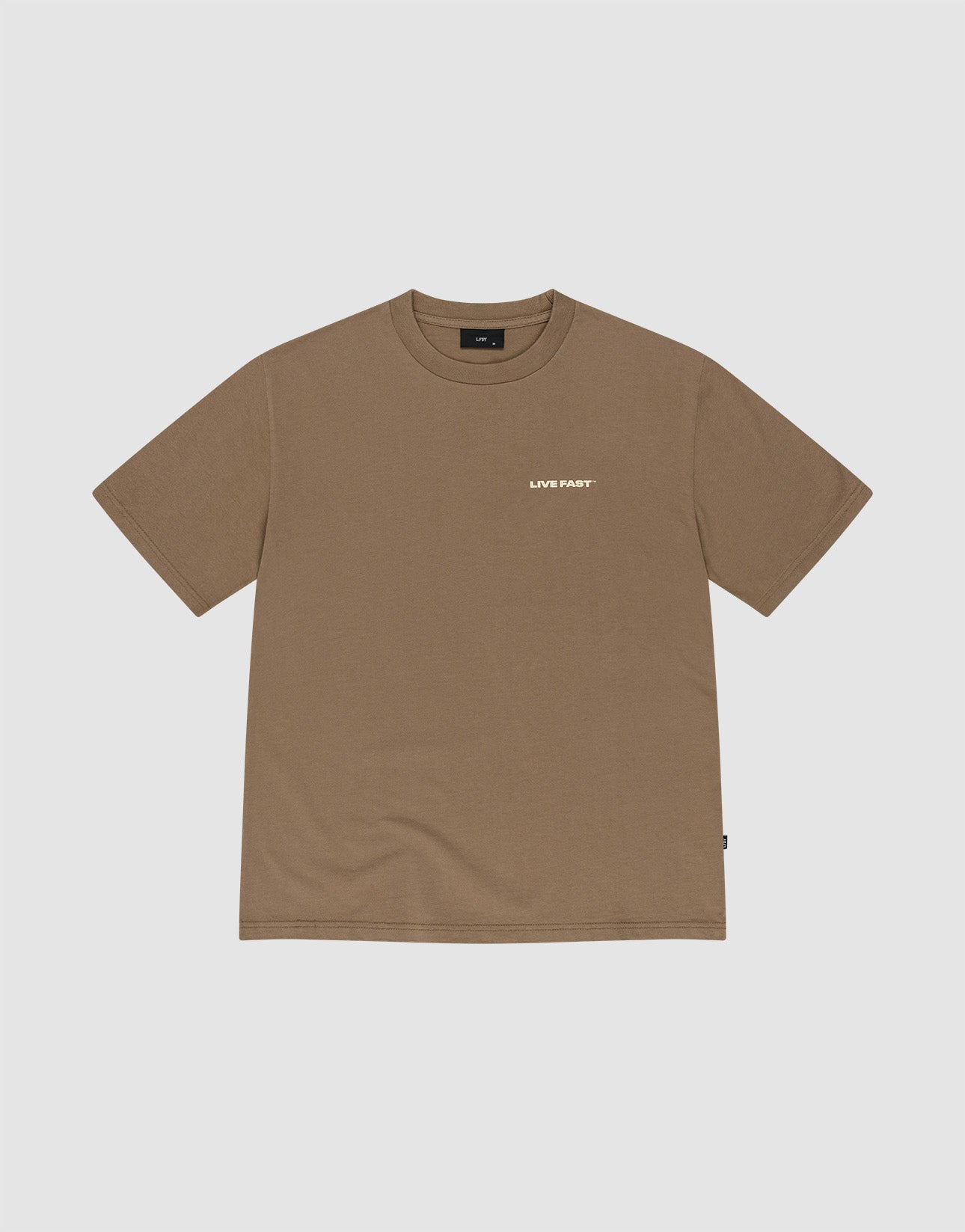 A young man with short dreadlocks wears the LIVE FAST DIE YOUNG Live Fast Plain Tee in brown (100% Baumwolle) featuring a subtle frontprint, paired with faded blue jeans. He stands against a light gray background, hands in his pockets.
