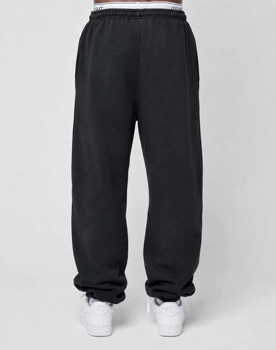 A person models the LIVE FAST DIE YOUNG Basic Loose Jogger in black, featuring an elastic waistband and subtle logo print, paired with white sneakers. The shot is from the back, focusing on the lower body against a plain background.