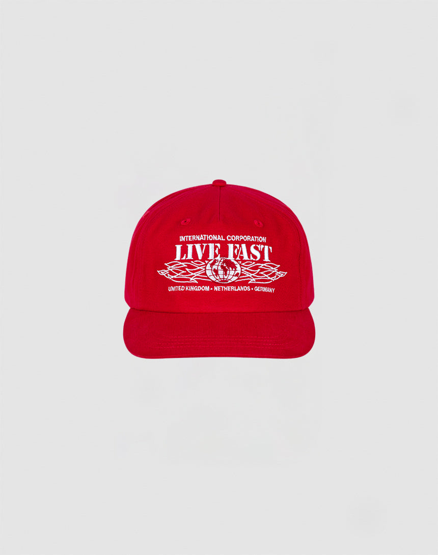 The Live Fast Corporation Cap by LIVE FAST DIE YOUNG is a 100% cotton red cap with bold white International Corporation LIVE FAST text and multi-logo graphics, plus country names United Kingdom, Netherlands, and Germany on the front.