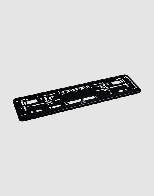 The LFDY License Plate Holder (2 Pack) by LIVE FAST DIE YOUNG is a black plastic Eurokennzeichen holder with multiple mounting slots and holes and a subtle LFDY logo, shown on a light gray background.| plain 